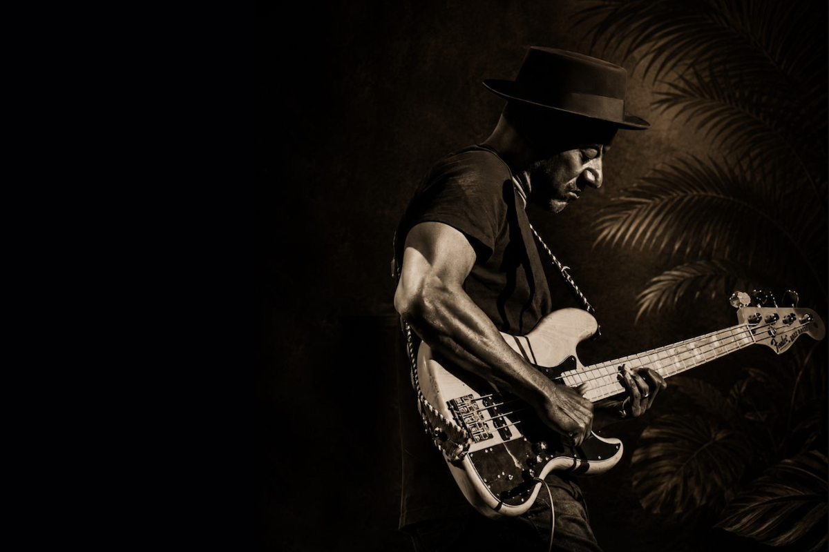 Jazz bassist Marcus Miller playing his Fender Jazz Bass guitar in a fedora hat against a moody, dark sepia background with palm trees.